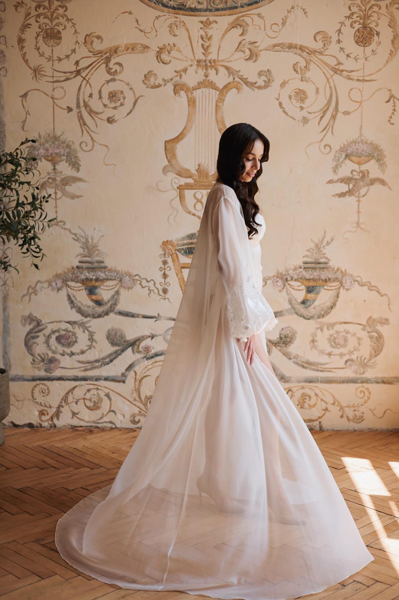 May include: A long, white sheer robe with sleeves and lace detailing. The robe is worn over a white garment. The background features a vintage-style wall mural with floral and scroll designs, and a wooden floor.