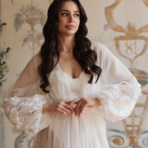 May include: A white, sheer robe with floral lace detailing on the sleeves. The robe is worn over a white dress. The background features a decorative wall with floral and architectural elements.