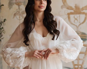 Puede incluir: Una bata blanca transparente con detalles de encaje floral en las mangas. La bata se usa sobre un vestido blanco. El fondo presenta una pared decorativa con elementos florales y arquitectónicos.