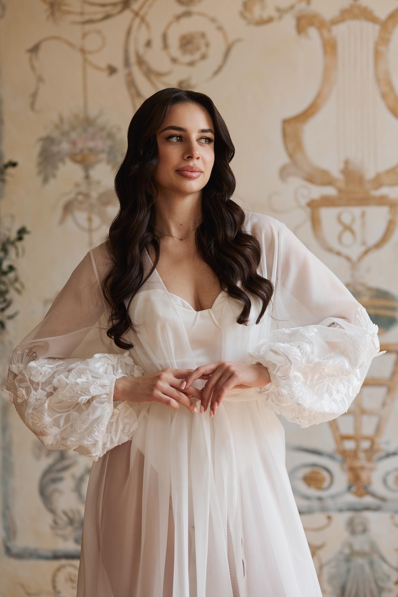 May include: A white, sheer robe with floral lace detailing on the sleeves. The robe is worn over a white dress. The background features a decorative wall with floral and architectural elements.