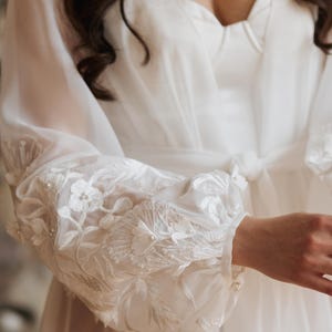 May include: A white bridal robe with sheer sleeves. The sleeves feature detailed floral embroidery and small pearl embellishments. The robe has a tie at the waist and a sweetheart neckline.