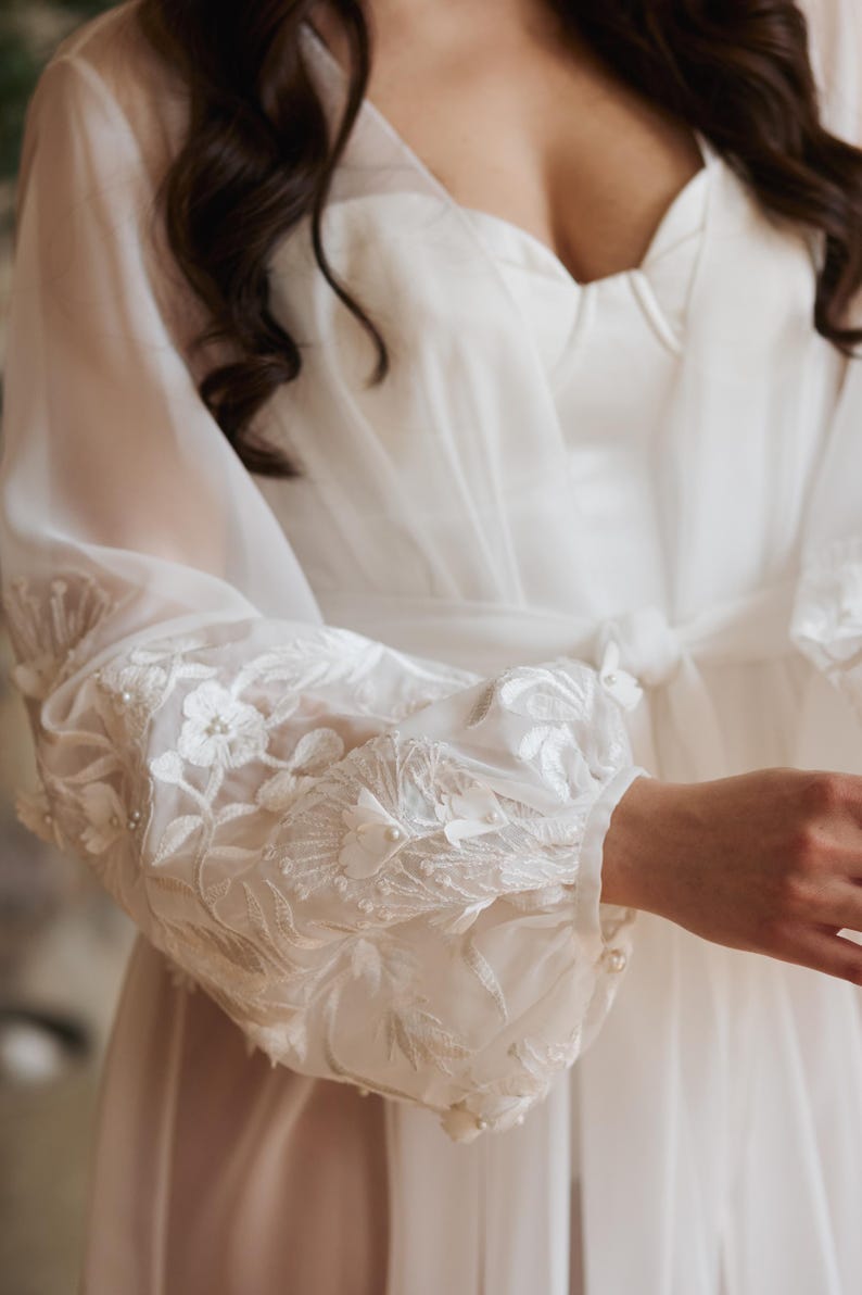May include: A white bridal robe with sheer sleeves. The sleeves feature detailed floral embroidery and small pearl embellishments. The robe has a tie at the waist and a sweetheart neckline.