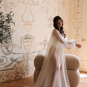 May include: A sheer white robe with lace sleeves and a flowing train. The robe is worn over a white garment. The background features a beige and cream-coloured wall with ornate painted details and a beige upholstered chair.