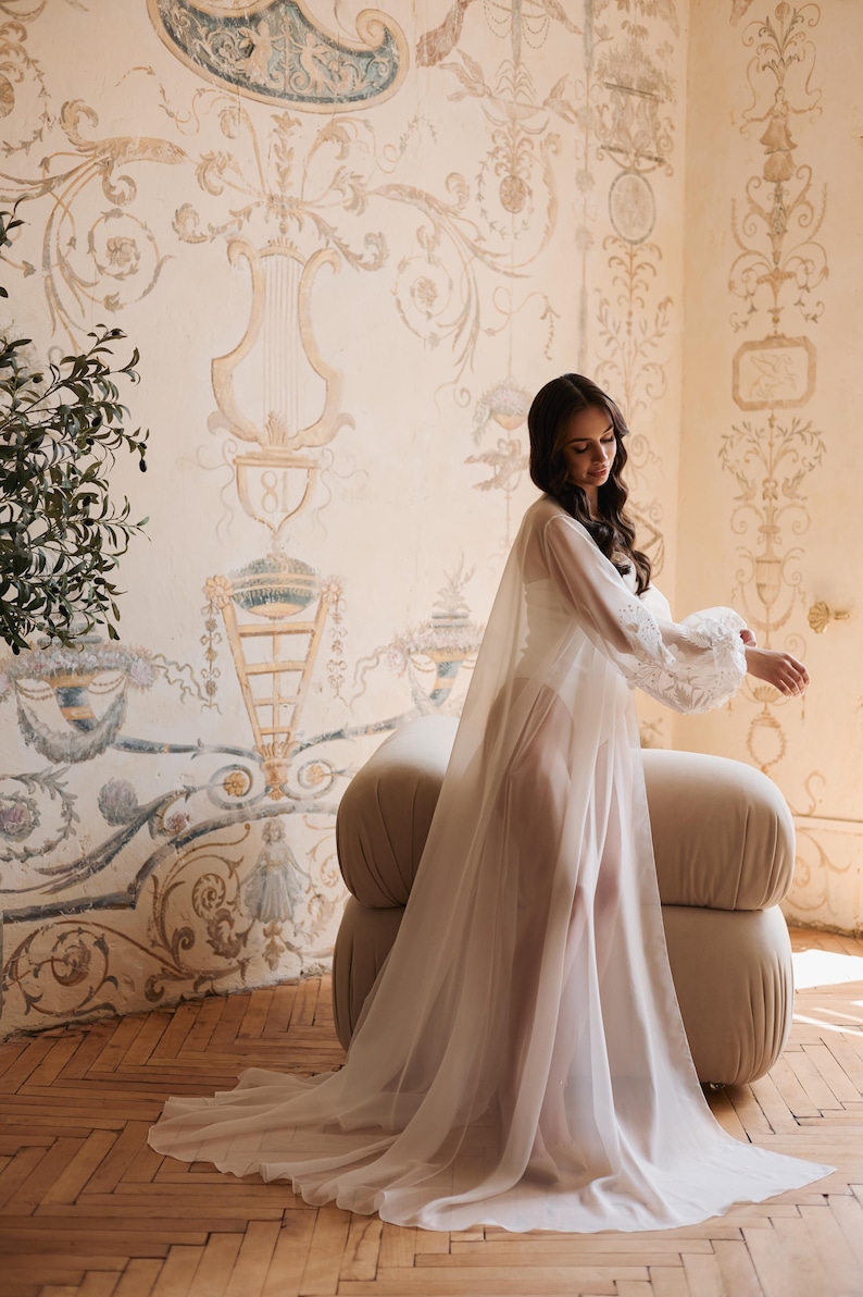 May include: A sheer white robe with lace sleeves and a flowing train. The robe is worn over a white garment. The background features a beige and cream-coloured wall with ornate painted details and a beige upholstered chair.