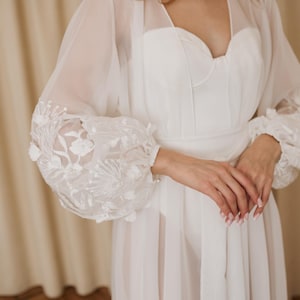 May include: A white bridal gown with a sweetheart neckline and sheer, long sleeves. The sleeves feature intricate floral embroidery at the cuffs. The dress appears to be made of a soft, flowing fabric, suitable for a wedding or special occasion.