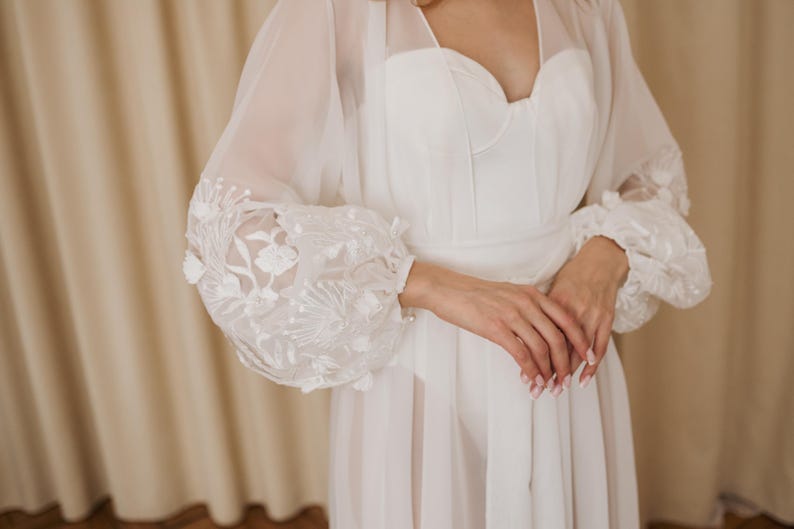 May include: A white bridal gown with a sweetheart neckline and sheer, long sleeves. The sleeves feature intricate floral embroidery at the cuffs. The dress appears to be made of a soft, flowing fabric, suitable for a wedding or special occasion.