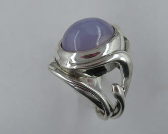 May include: A silver ring with a large, oval-shaped, lavender gemstone set in a smooth, polished bezel. The ring band is intricately designed with a swirling, organic pattern.