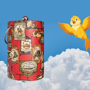 May include: A red and white cylindrical ice bucket with a silver lid and a silver handle. The bucket is covered in a pattern of vintage illustrations. A yellow bird with orange wings is flying in the sky above a white cloud.