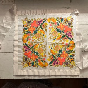 May include: A white fabric chair cushion with a floral design in shades of orange, yellow, green, and pink. The cushion has a white ruffle trim around the edges.