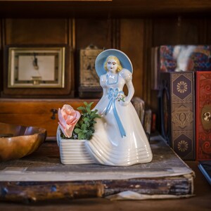 May include: A white ceramic figurine of a woman wearing a blue dress and a hat. She is holding a bouquet of flowers and standing on a white ceramic base. The figurine is on a wooden shelf with other decorative items.