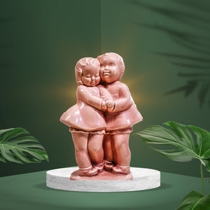 May include: A pink ceramic figurine of two children hugging each other. The figurine is on a white pedestal.