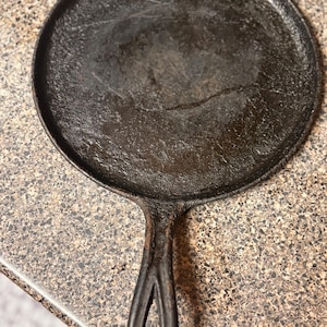 May include: A well-seasoned, round cast iron griddle with a single handle. The griddle is dark gray with a slightly textured surface and a raised edge. The handle has a teardrop-shaped opening.