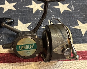 May include: A dark gray metal fishing reel with a black handle. The side plate features a circular label with the text MODEL 870B, LANGLEY, SPINATOR, and MADE IN U.S.A. The reel has a metallic bail arm and a textured finish on the main body housing.
