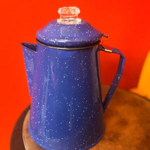 May include: A blue speckled enamelware coffee pot with a clear glass knob on the lid. The pot has a handle and a spout. The background is a solid red color. The coffee pot is sitting on a wooden surface.