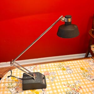 May include: A black desk lamp with a silver adjustable arm and a black lampshade. The lamp sits on a dark gray base. The background is a solid red wall. The lamp is on a yellow and white checkered tablecloth with a floral and bunny pattern.