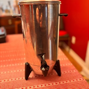 May include: A vintage stainless steel coffee urn with a glass lid and black plastic accents. The urn has a spigot and sits on three black legs. It is on a red and white checkered tablecloth.
