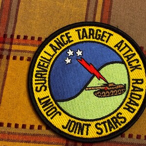 May include: Circular patch with the words "SURVEILLANCE TARGET ATTACK RADAR JOINT STARS" in yellow. The patch features a blue sky with three white stars, a green landscape, and a red lightning bolt over a tank.