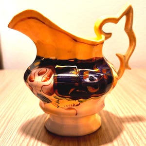 May include: A decorative ceramic pitcher with a yellow rim and handle, and a base in a cream color. The body of the pitcher is decorated with a dark blue and orange floral pattern. The pitcher has a unique handle design.