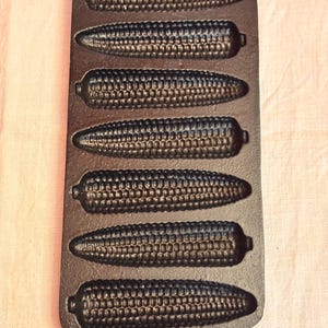 May include: A cast iron cornbread pan with six corn cob-shaped molds. The pan is dark brown and has a loop at the top for hanging. The molds are detailed to resemble corn kernels.