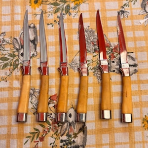 May include: Set of six steak knives with silver blades and red handles. The handles are a light tan color with silver accents at the base. The knives are arranged on a yellow and white checkered tablecloth with floral and animal patterns.