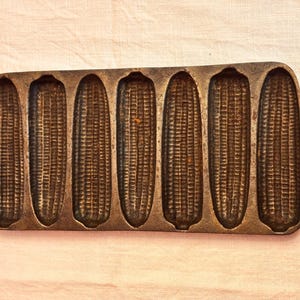 May include: A cast iron cornbread pan with six corn-shaped molds. The pan is dark gray with handles on each end. The molds are detailed to resemble corn on the cob. The pan is designed for baking cornbread in the shape of corn.