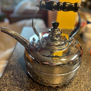 May include: A polished chrome tea kettle featuring a rounded body, a curved spout, and a black wooden handle with a decorative turned shape. The lid includes a small black knob, and the base has a perforated metal design for heat distribution.