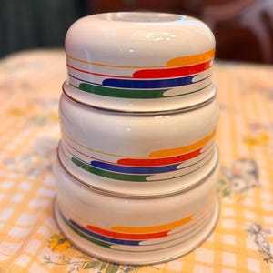 May include: Stacked set of three white mixing bowls with a retro design. Each bowl features a colorful horizontal stripe pattern in orange, red, blue, and green. The bowls are made of a shiny, reflective material and have a silver rim.