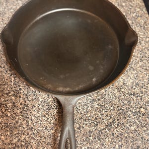May include: A well-seasoned cast iron skillet with a dark, matte finish. The pan has a long handle with a hole for hanging and two small spouts. The skillet is designed for cooking and is likely used for frying, searing, and baking.