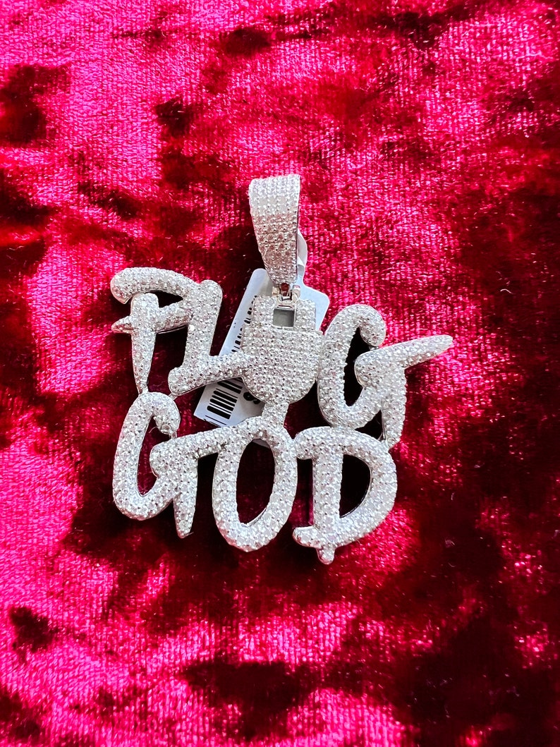 May include: A silver pendant with the words "Plug God" set in sparkling white stones. The pendant is on a red velvet background.