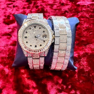 May include: A luxury watch and matching bracelet set. The watch features a round face with a diamond-like encrusted bezel and dial. The watch band and bracelet are also encrusted with similar stones. The set is displayed on a blue pillow against a red velvet background.