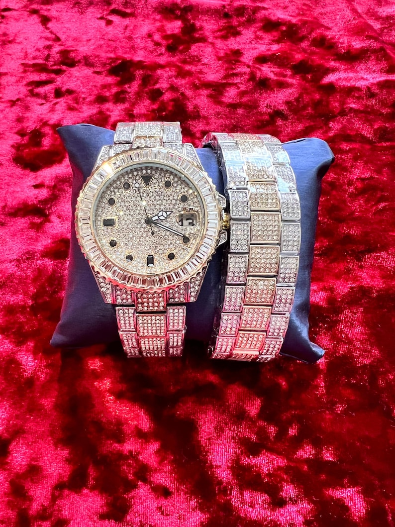 May include: A luxury watch and matching bracelet set. The watch features a round face with a diamond-like encrusted bezel and dial. The watch band and bracelet are also encrusted with similar stones. The set is displayed on a blue pillow against a red velvet background.
