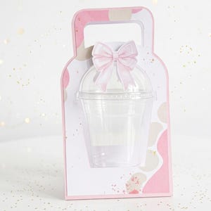 May include: A pink and white gift box with a handle, featuring a clear plastic cup and a pink bow. The box has a patterned design with pink and beige shapes. The bow is made of pink ribbon.