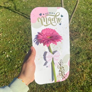 Puede incluir: Una tarjeta de felicitación con un diseño rosa y blanco, con una margarita rosa y el texto "Feliz Dia de la Madre". La tarjeta tiene un lazo rosa y un tallo verde. El fondo es una zona de césped.