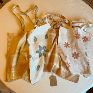 May include: Four fabric tote bags with handles. One is solid yellow-gold with tie-dye patterns. Another is off-white with green leaf prints. Two are off-white with rust-colored flower prints and tie-dye accents. A small brown tag reads Wild Oak Crafts.