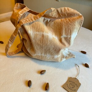 May include: A tan-colored tote bag with a tie-dye pattern and long straps. The bag is made of fabric and has a tag that reads "Wild Color Crafts". Several brown acorns are scattered around the bag on a white surface.