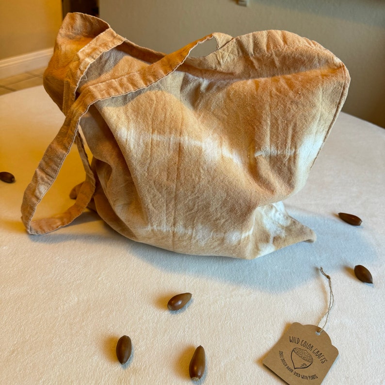 May include: A tan-colored tote bag with a tie-dye pattern and long straps. The bag is made of fabric and has a tag that reads "Wild Color Crafts". Several brown acorns are scattered around the bag on a white surface.