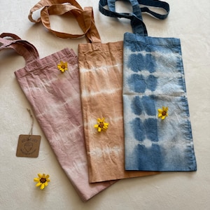May include: Three tie-dye tote bags with long straps. The bags are in shades of pink, orange, and blue. Each bag has a different tie-dye pattern. The bags are on a white surface with small yellow flowers.