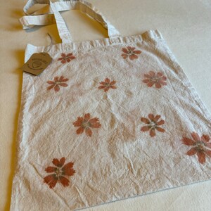 May include: Off-white tote bag with a repeating pattern of reddish-brown floral designs. The bag has matching off-white handles and a small tag attached. The bag is made of a textured fabric.