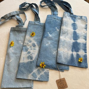 May include: Four blue and white tie-dye tote bags with yellow flowers on top of them. The bags have long straps and are made of a soft, natural fabric. The bags are arranged in a row on a white surface. A small brown tag with the text "LILA GOMEZ CRAFTS" is in the bottom right corner of the image.