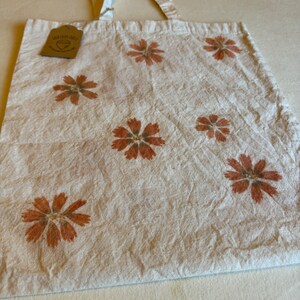 May include: Cream-colored tote bag with a floral pattern. The bag features several brown flower designs. The bag has a tag that reads "Wild Earth Lab Co.". The bag has a simple design and is made of a natural-looking fabric.