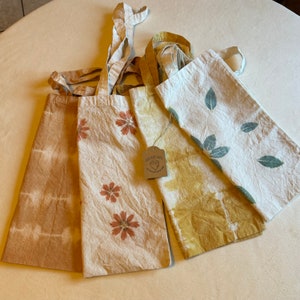 May include: Four eco-friendly tote bags with unique designs. One bag features brown tie-dye, another has orange flowers, a third has yellow and brown patterns, and the last has green leaves. Each bag has a natural, textured appearance.