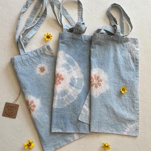 May include: Three blue tote bags with tie-dye patterns in shades of pink and white. The bags have long straps and are made of a soft, natural fabric.