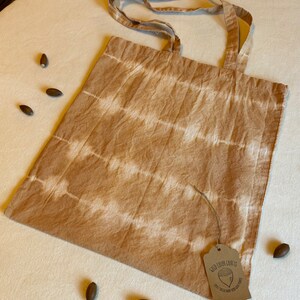 May include: A beige tote bag with horizontal white stripes. The bag has matching handles and a small tag with text and a graphic. Several acorns are scattered around the bag on a white surface.