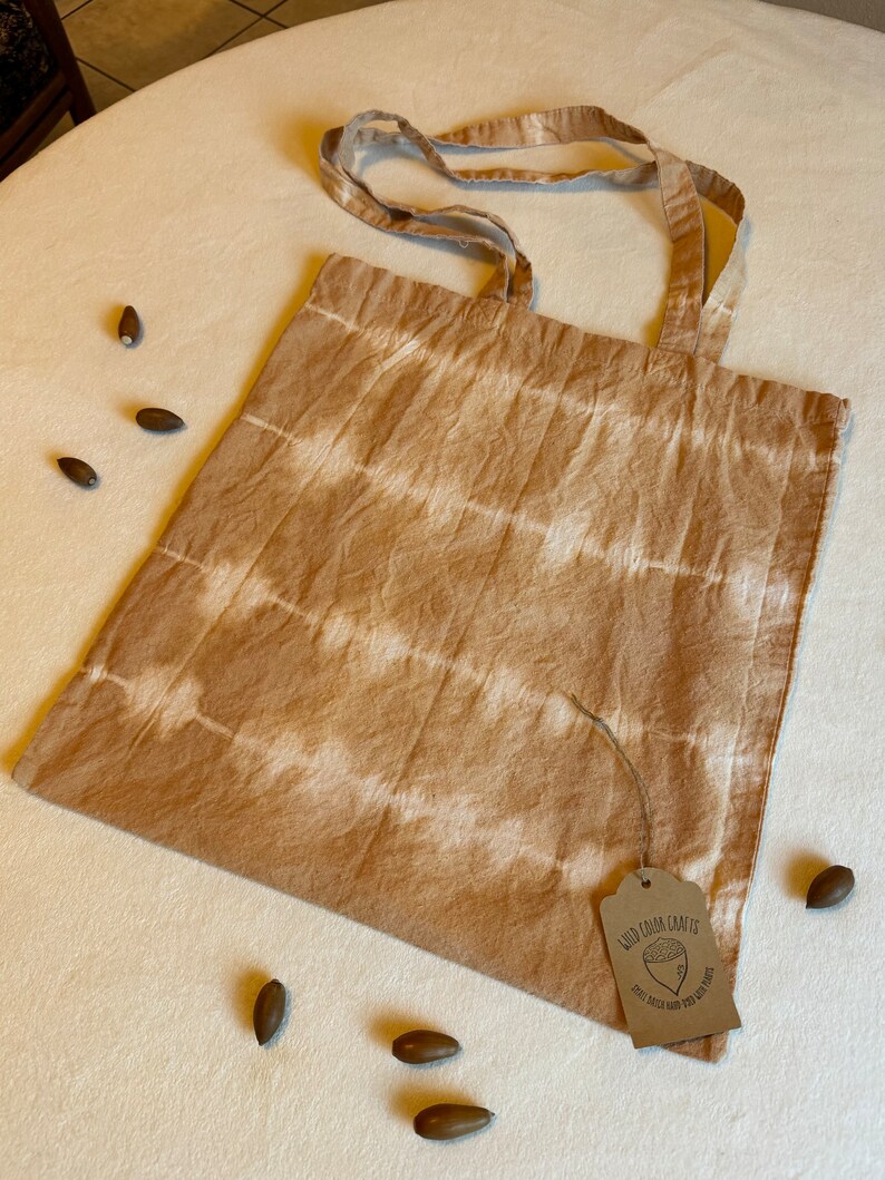 May include: A beige tote bag with horizontal white stripes. The bag has matching handles and a small tag with text and a graphic. Several acorns are scattered around the bag on a white surface.