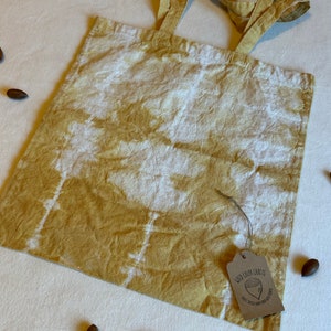 May include: A mustard yellow and white tie-dye tote bag with matching handles. A brown tag with the text "Wild Color Crafts" and a small acorn illustration is attached to the bag. Several acorns are scattered around the bag.