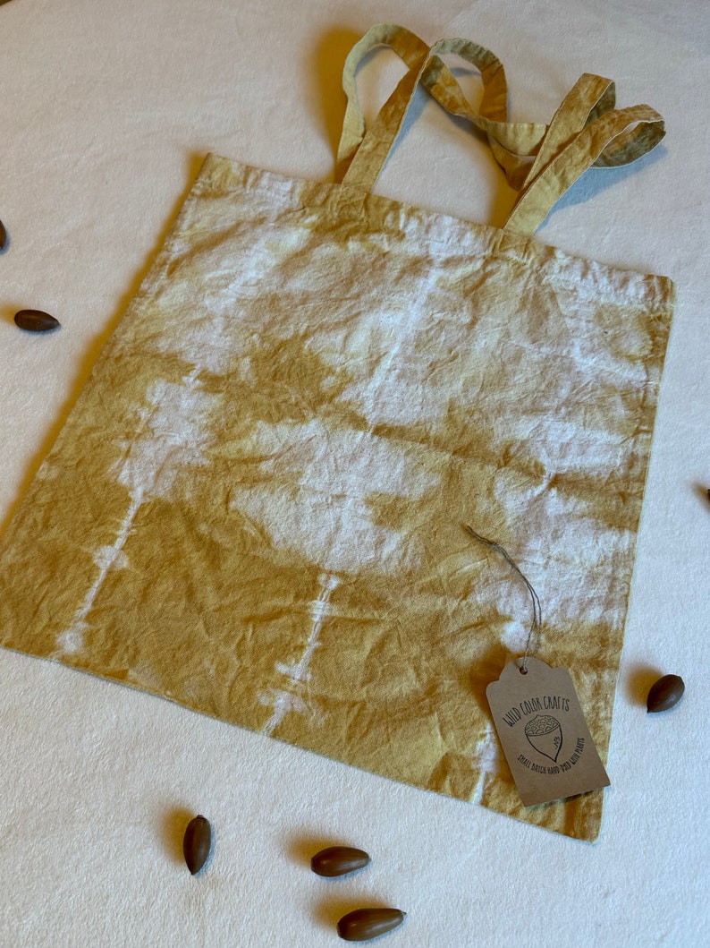 May include: A mustard yellow and white tie-dye tote bag with matching handles. A brown tag with the text "Wild Color Crafts" and a small acorn illustration is attached to the bag. Several acorns are scattered around the bag.