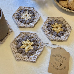 May include: Three crocheted hexagon coasters in beige with a brown and yellow flower design. The coasters are on a white surface with a brown tag that reads "Wild Color Crafts, Small Batch Hand-Dyed with Plants".