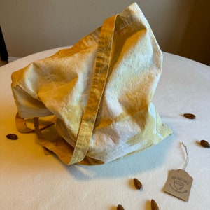 May include: A handmade tote bag with a natural, organic look. The bag is a light yellow color with darker yellow accents and long straps. The tag reads "Wild Color Crafts". Several acorns are scattered around the bag.