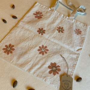 May include: A cream-colored tote bag with a floral pattern in shades of brown. The bag has long handles and a tag that reads "Wild Color Crafts". Several acorns are scattered around the bag.