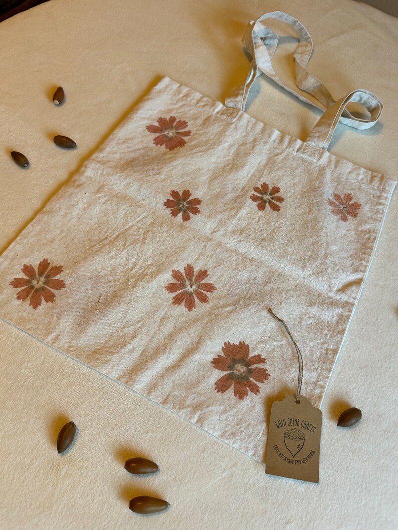 May include: A cream-colored tote bag with a floral pattern in shades of brown. The bag has long handles and a tag that reads "Wild Color Crafts". Several acorns are scattered around the bag.